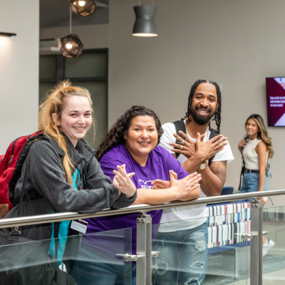 Three female students and one male student with dreadlocks pose with the Riverbat sign against a metal and glass rail at ACC Highland campus.