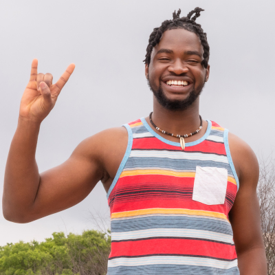 A smiling young man wearing a colorful striped tank top, standing outdoors and making the “Hook ’em Horns” hand sign, a symbol associated with the University of Texas at Austin. The image conveys pride and enthusiasm for the PACE co-enrollment program with UT Austin.