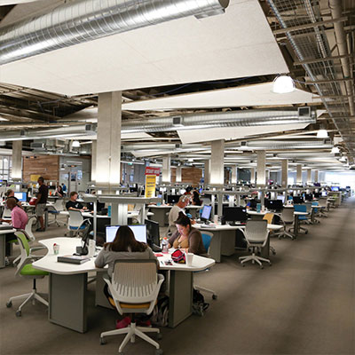 A large, open workspace filled with students sitting at desks with computers, working individually and in small groups. The room has modern lighting and exposed ductwork, giving it a contemporary, industrial feel.