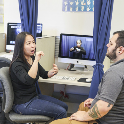 A woman and a man practicing American Sign Language (ASL) in a classroom with computer monitors showing ASL videos in the background. The woman is signing, and the man is attentively watching, emphasizing the interactive nature of interpreter training.